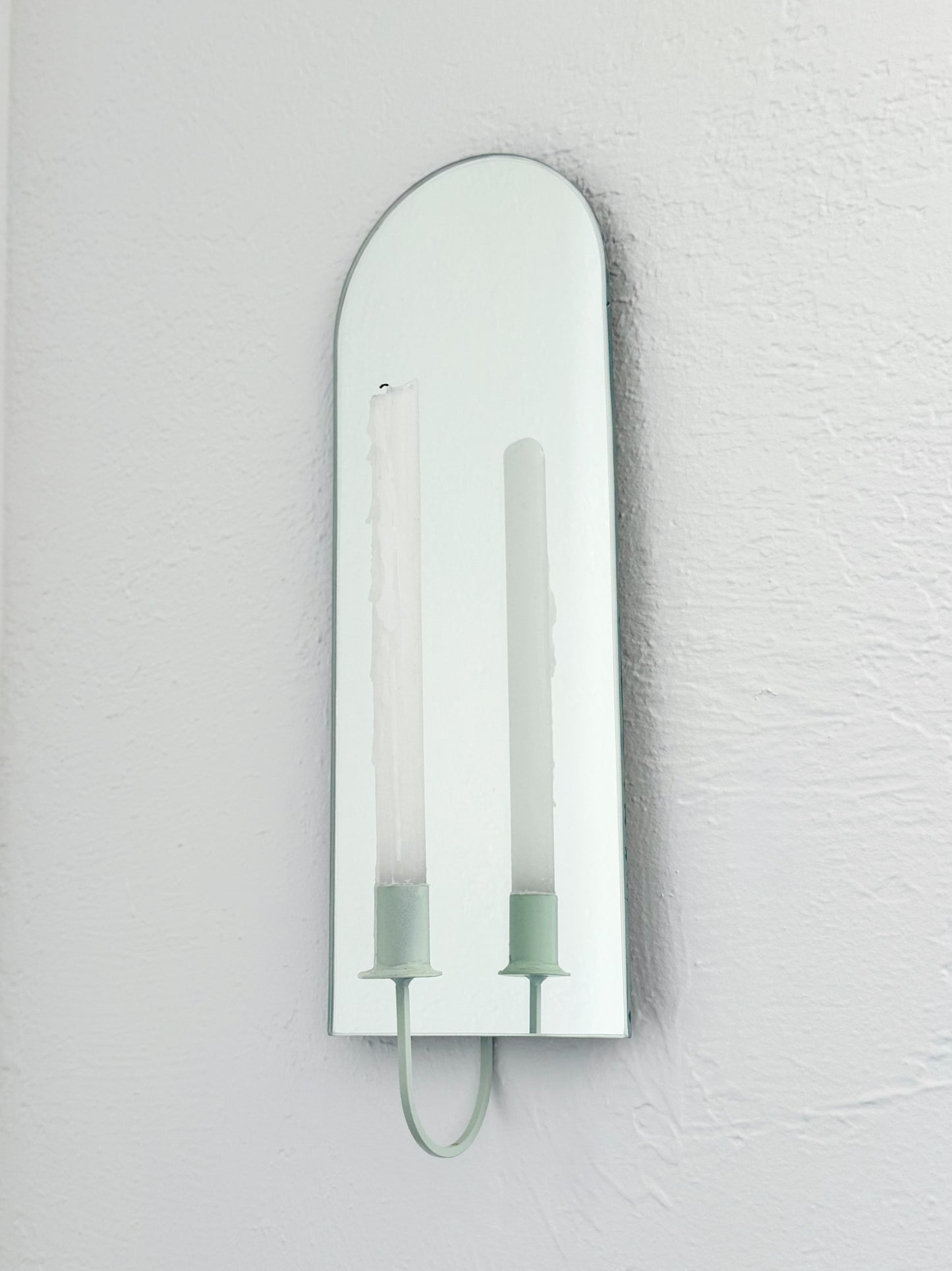 The Arch Mirror Candle Sconce or Mirror Candle Holder is wall mounted to a white wall. The candleholder is light green & holding a white unlit candle. The reflection in the mirror is only of the light green candle holder & the tall white candle.
