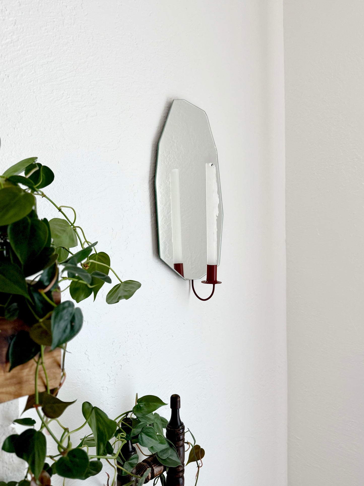 Side angle of Gem Cut Mirror Candle Sconce or Mirror Candle Holder is wall mounted. The candleholder is deep red, holding a white unlit candle. The reflection in the mirror is of the red candle holder & candle. The setting is a cozy home with plants.