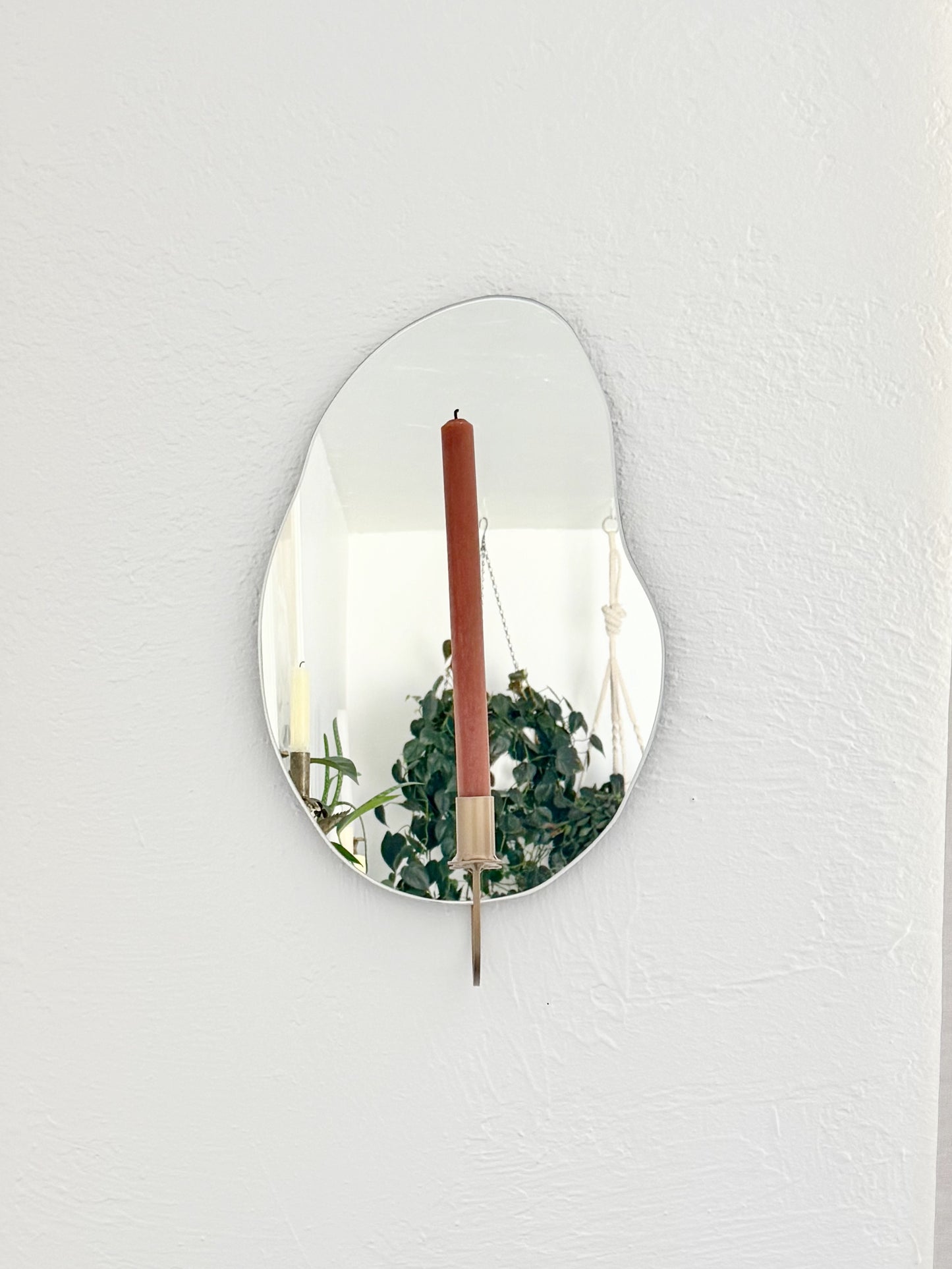 Straight shot of the wall mounted organic shape Mar Mirror Candle Sconce or Mirror Candle Holder. The candleholder is gold & holding a red brown unlit candle. The reflection is of the gold candle holder, the unlit candle & the cream background with hanging plants & decor.