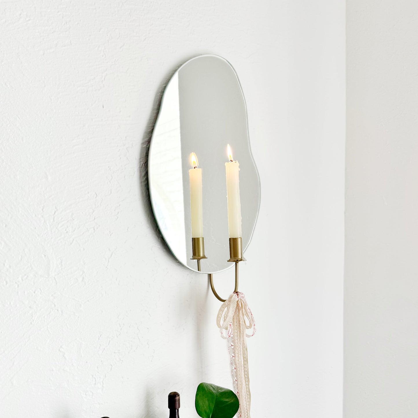 Side angle of the wall mounted organic shape Mar Mirror Candle Sconce or Mirror Candle Holder. The candleholder is gold with lacy ribbon tied to the base to give a whimsical aesthetic. The reflection is of the cream lit candle & gold candle holder. The setting is in a home with plants & a vintage chair.