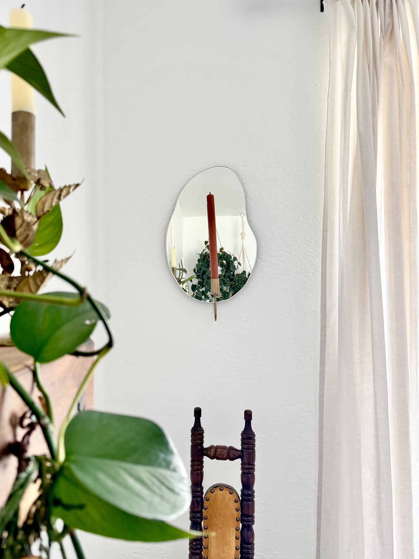 Straight shot of the wall mounted organic shape Mar Mirror Candle Sconce or Mirror Candle Holder. The candleholder is gold & holding a red brown unlit candle. The reflection is of the gold candle holder, the unlit candle & the cream background with hanging plants & decor. The setting is a cozy home with a vintage chair and plants.
