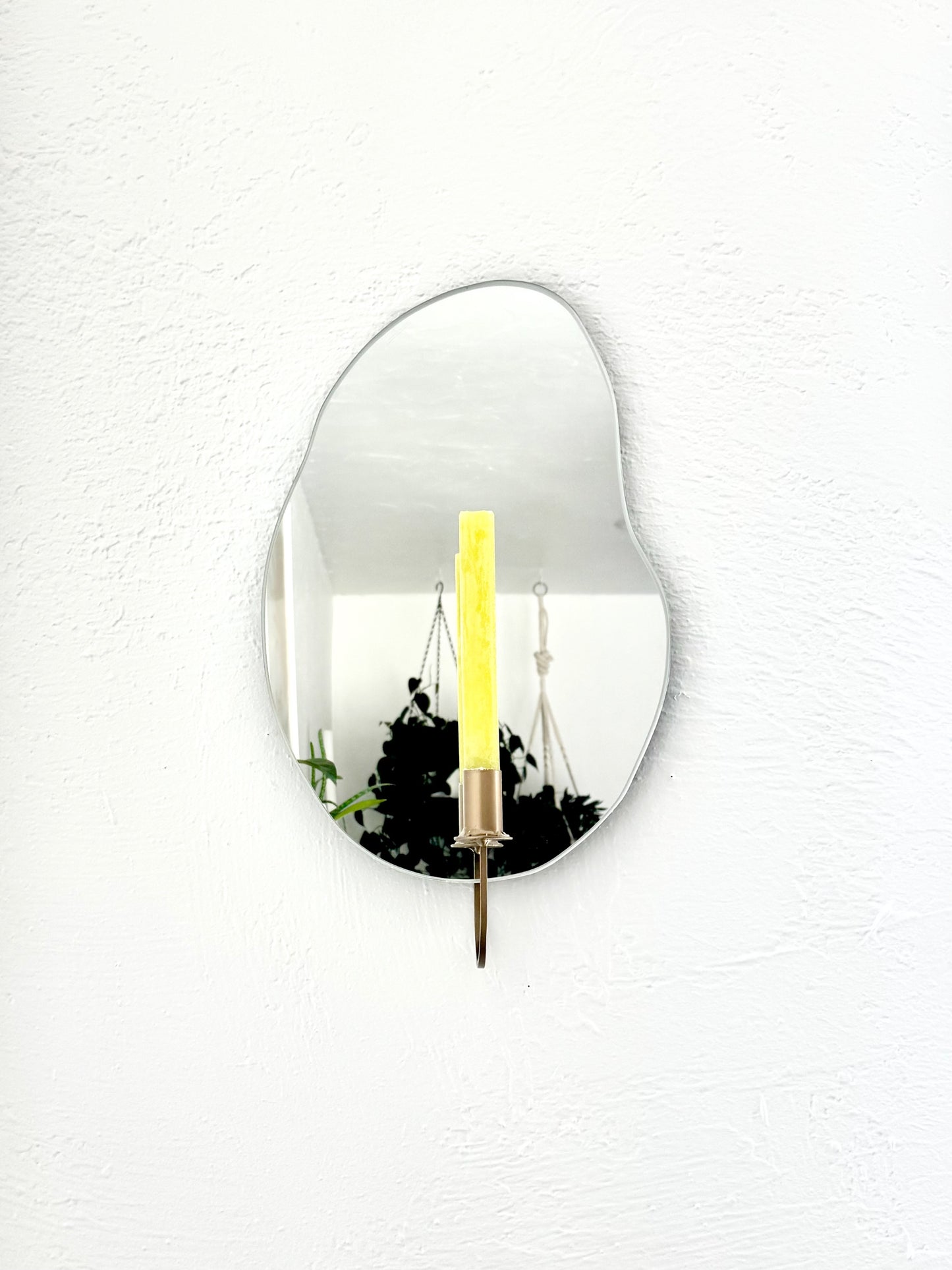 Straight shot of the wall mounted organic shape Mar Mirror Candle Sconce or Mirror Candle Holder. It has a gold candleholder & holding a green unlit candle. The Mirror Candle Holder is in a cozy home setting. The reflection is of the gold candle holder, the green unlit candle & hanging plants.