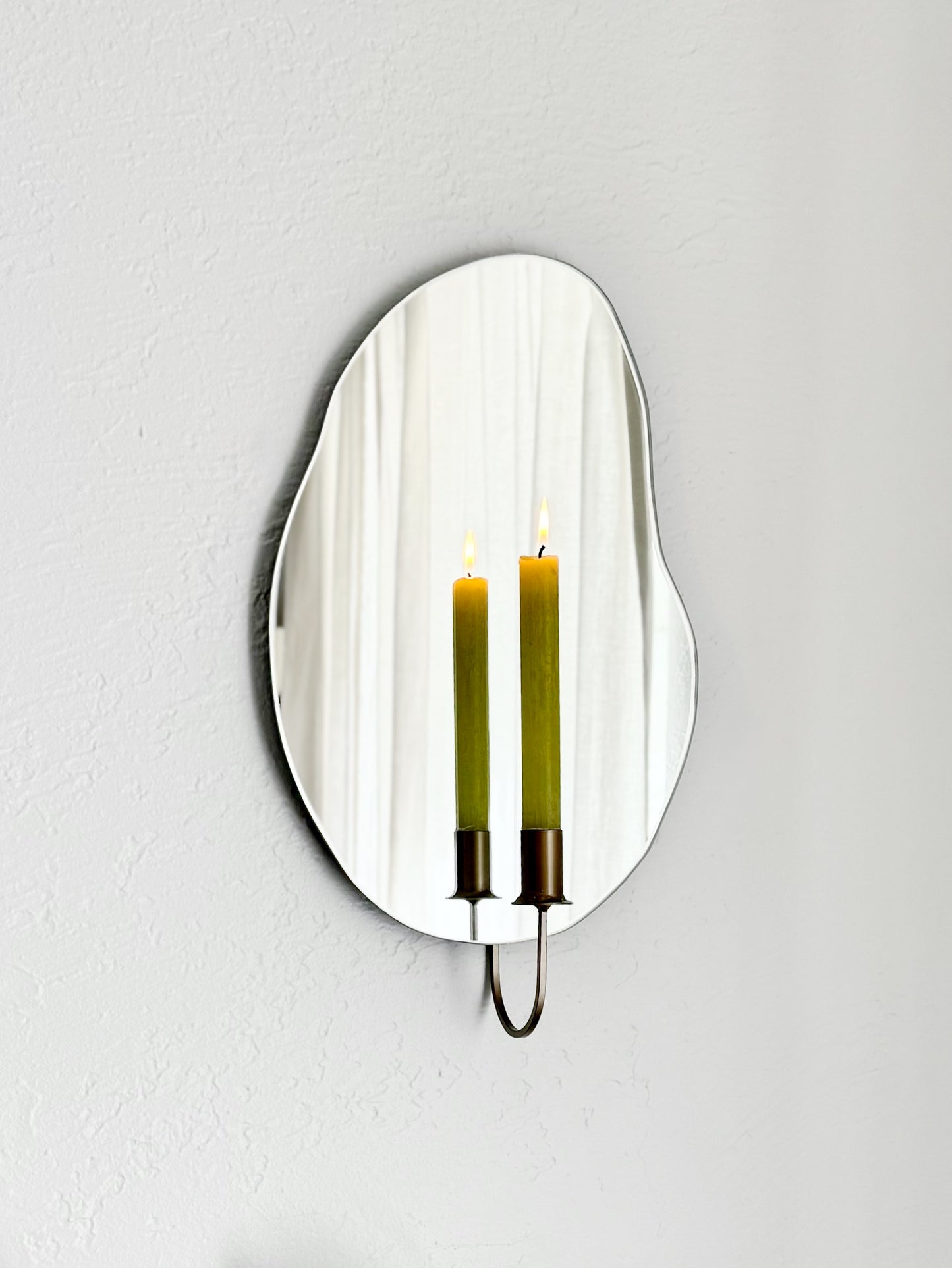 Angled shot of the wall mounted organic shape Mar Mirror Candle Sconce or Mirror Candle Holder. It has a gold candleholder & is holding a green lit candle. The reflection is of the gold candle holder, the green lit candle & the cream background.