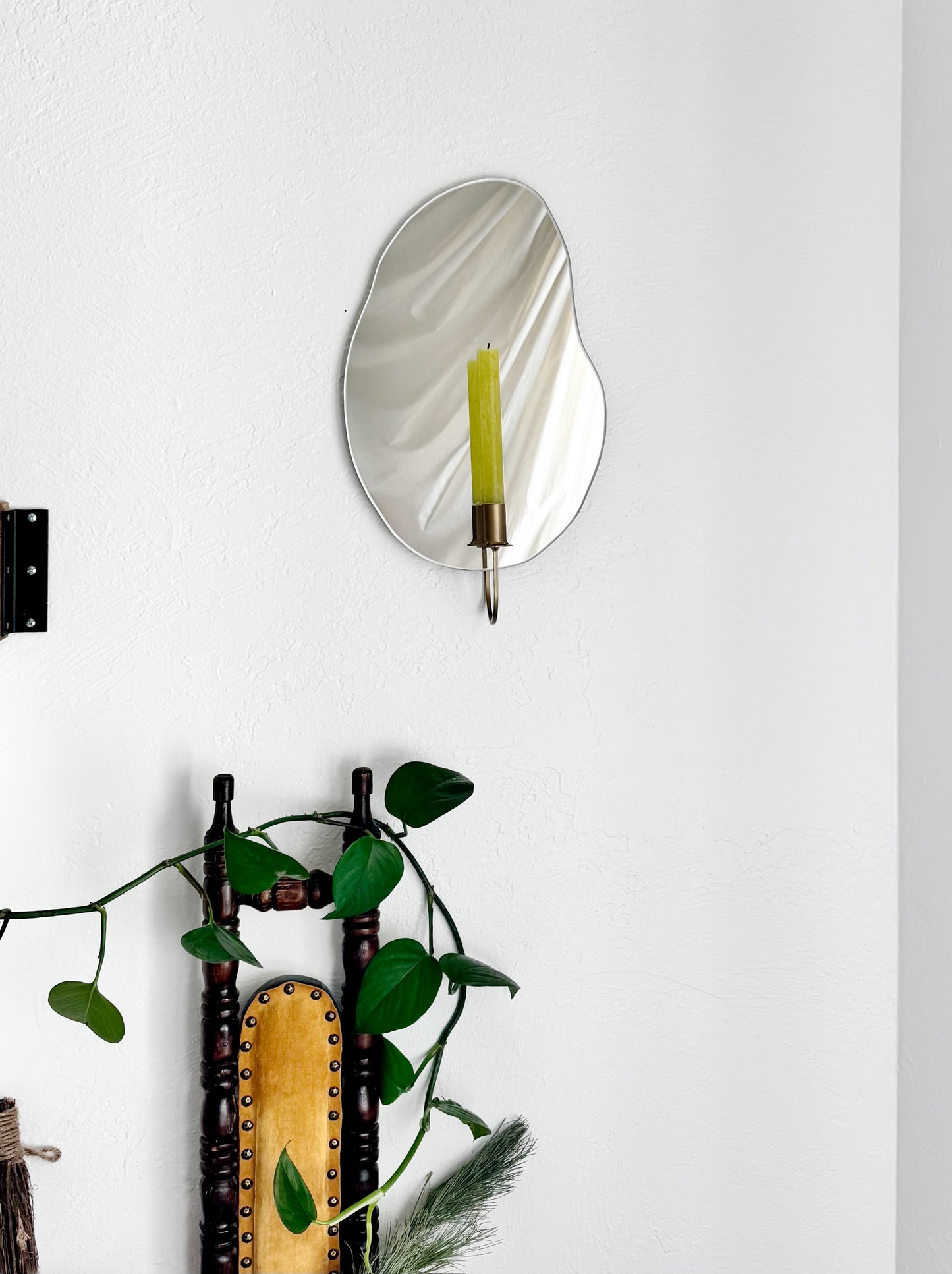 Straight shot of the wall mounted organic shape Mar Mirror Candle Sconce or Mirror Candle Holder. It has a gold candleholder holding a green unlit candle. The reflection is of the gold candle holder, the green unlit candle & the cream background. It is a home setting with plants and a vintage wood chair with golden yellow cushions.