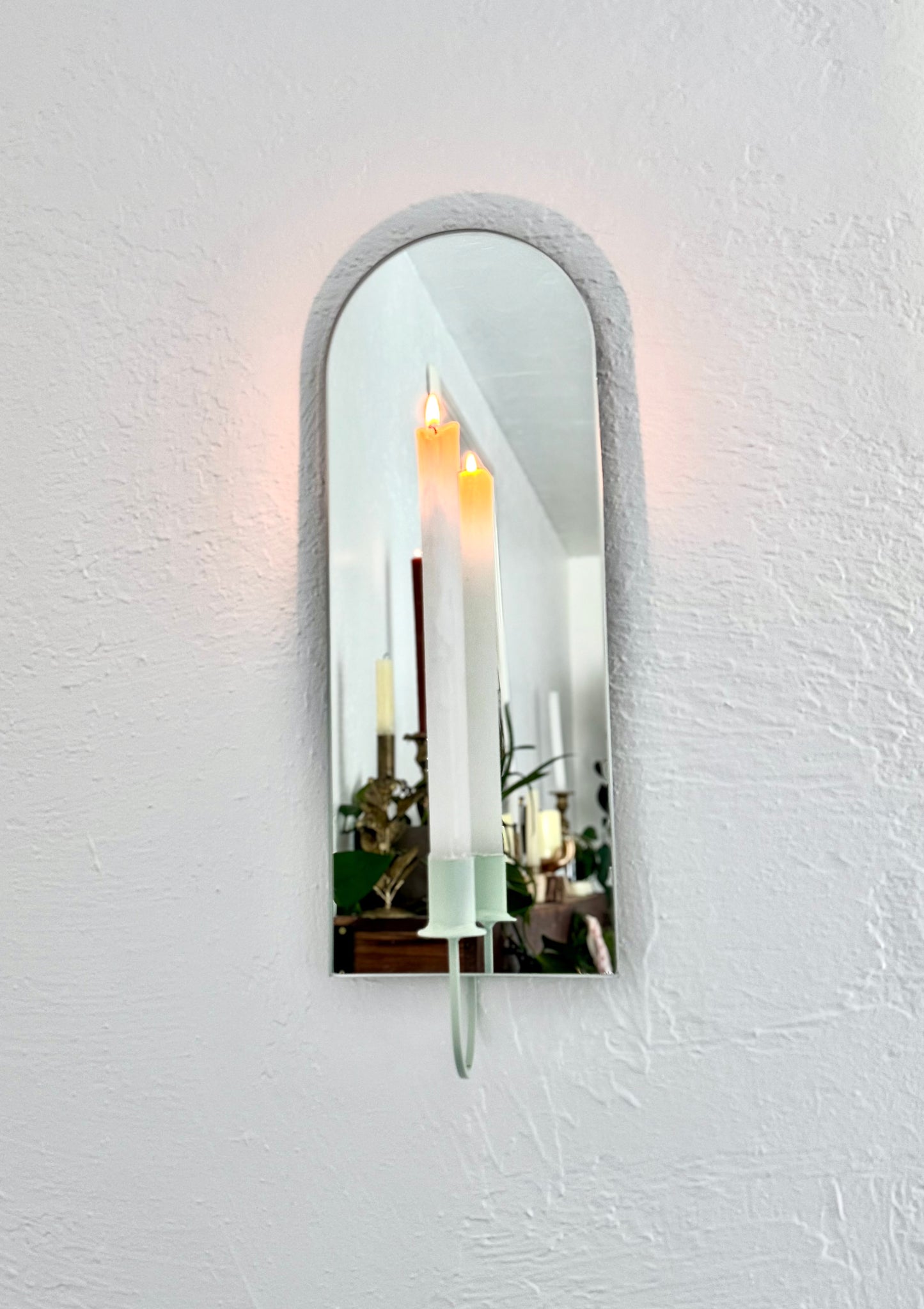 The Arch Mirror Candle Sconce or Mirror Candle Holder is wall mounted. The candleholder is light green & holding a white lit candle. The reflection is of the light green candle holder with the lit candle in a cozy home setting with plants and decor.