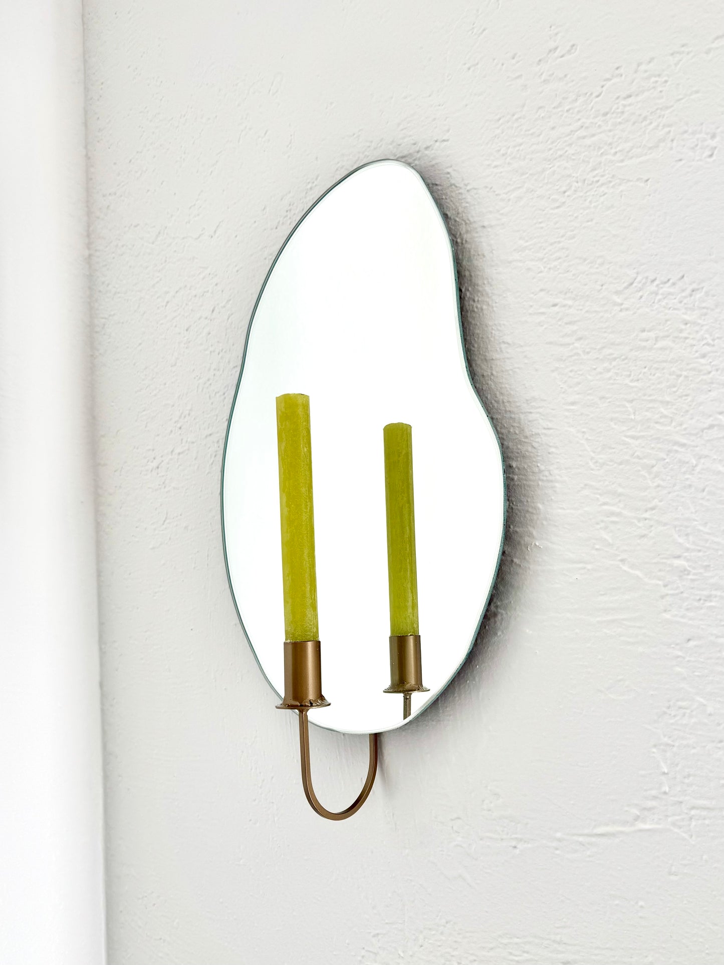 Side angle of the wall mounted organic shape Mar Mirror Candle Sconce or Mirror Candle Holder. The candleholder is gold & holding a green unlit candle. The reflection is of the gold candle holder, the green unlit candle & the white background.