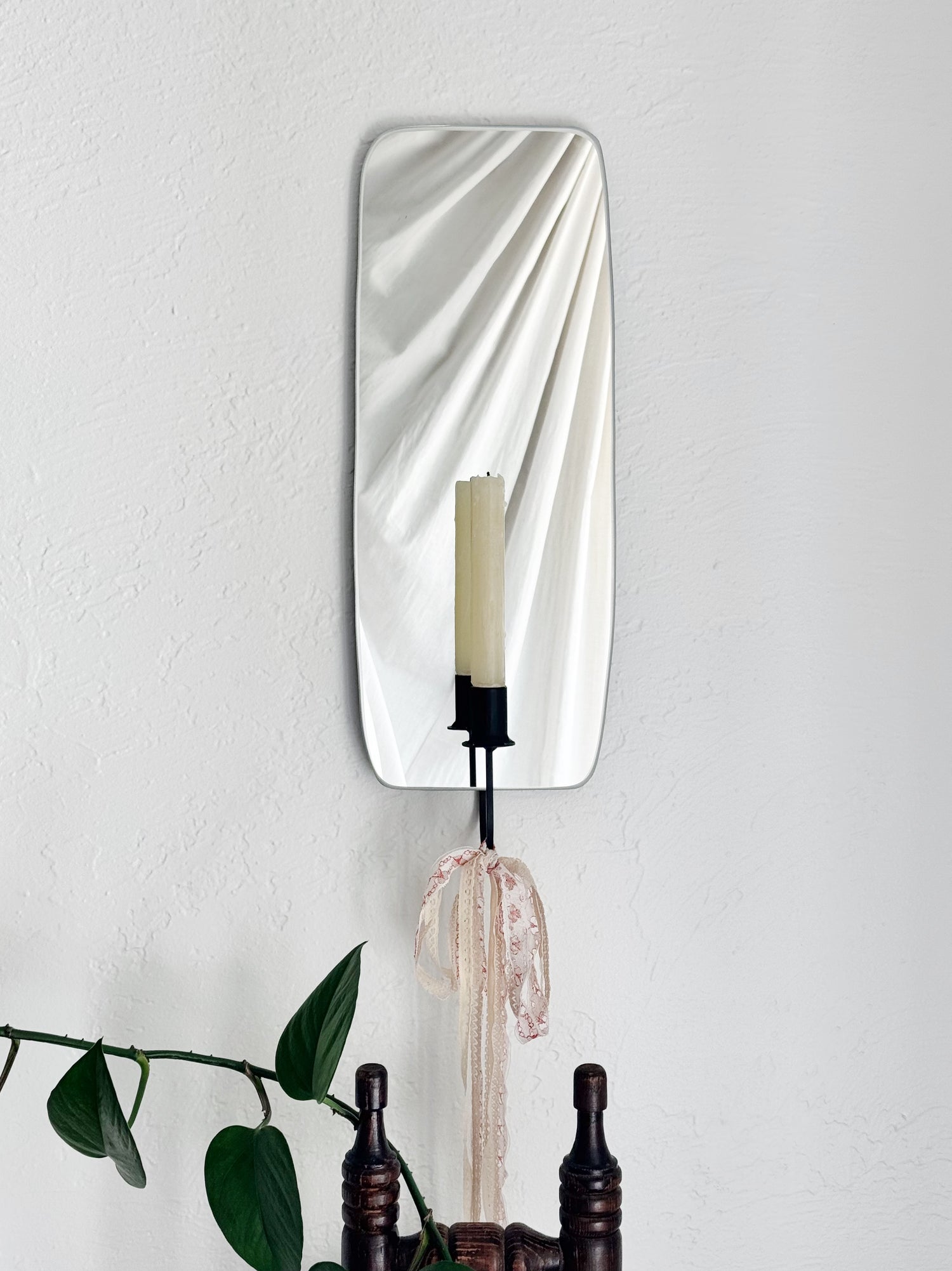 Mirror Candle Sconce or Mirror Candle Holder wall mounted. The black candleholder has lacy pink & cream ribbons tied to bottom to create a whimsical aesthetic. Reflection is of the candle holder, a cream unlit candle, plants & cozy decor.