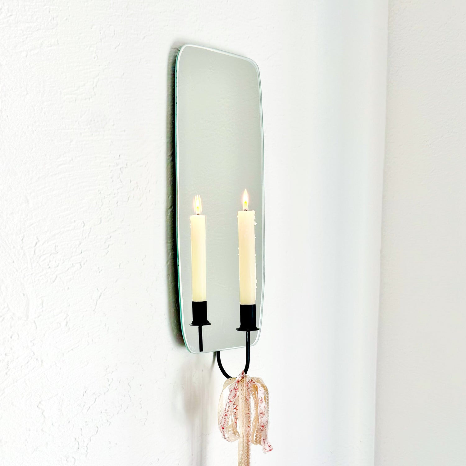 Side angle of the Oar Mirror Candle Sconce or Mirror Candle Holder wall mounted. The black candleholder has lacy pink & cream ribbons tied to bottom to create a whimsical aesthetic. The reflection is of the candle holder & a cream lit candle with the flame reflected giving it a cozy home feeling.