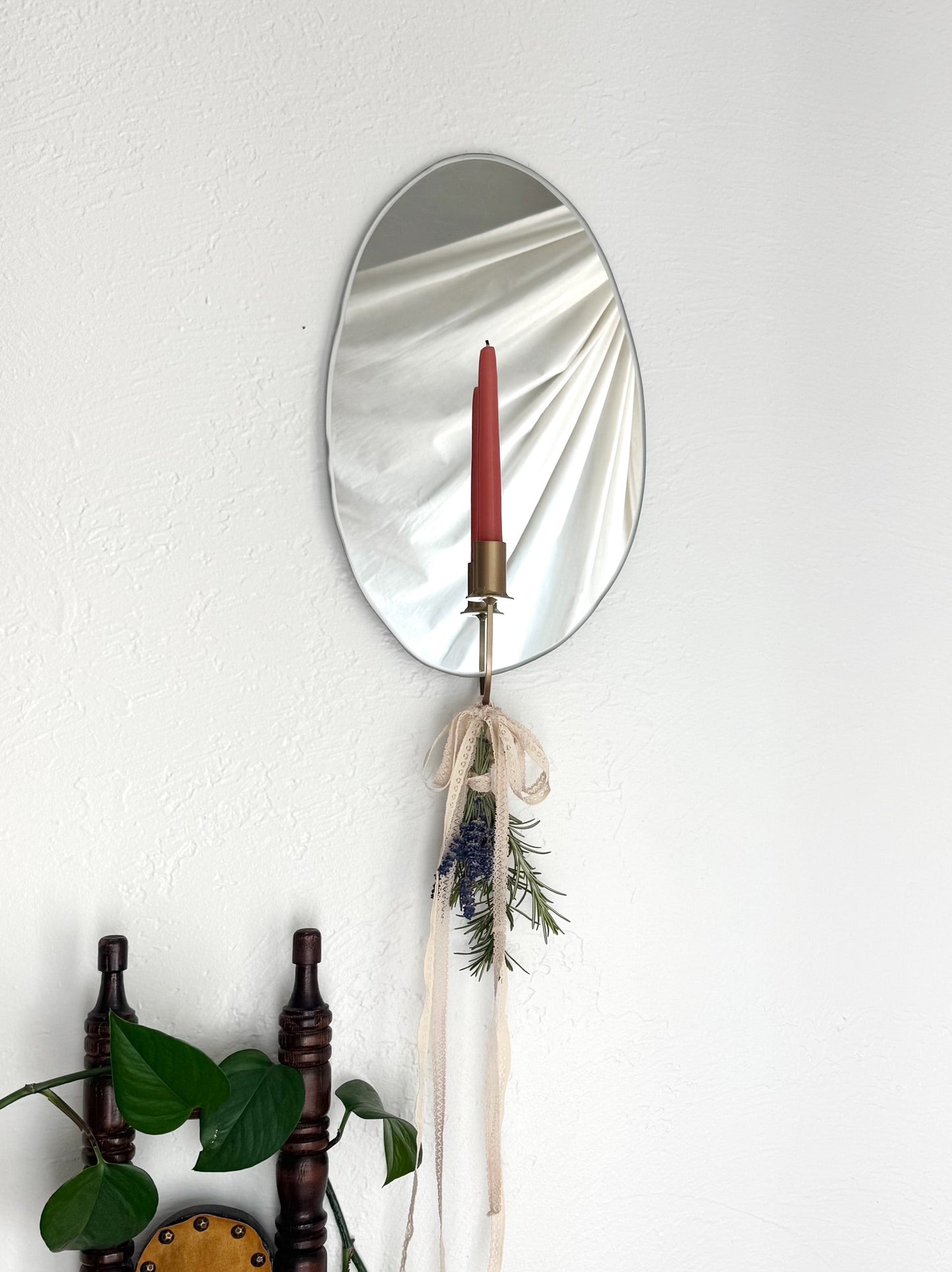 Straight view of the Mirror Candle Sconce or Mirror Candle Holder, wall mounted, with the gold candleholder and a red candle. The base of the gold candle holder has cream ribbon tied into a bow with a small bundle of dried herbs (rosemary & lavender) creating an inviting aesthetic. The reflection shows the light backdrop. In the foreground is the top of a small vintage wooden chair with an ivy plant hanging from the back of the chair.