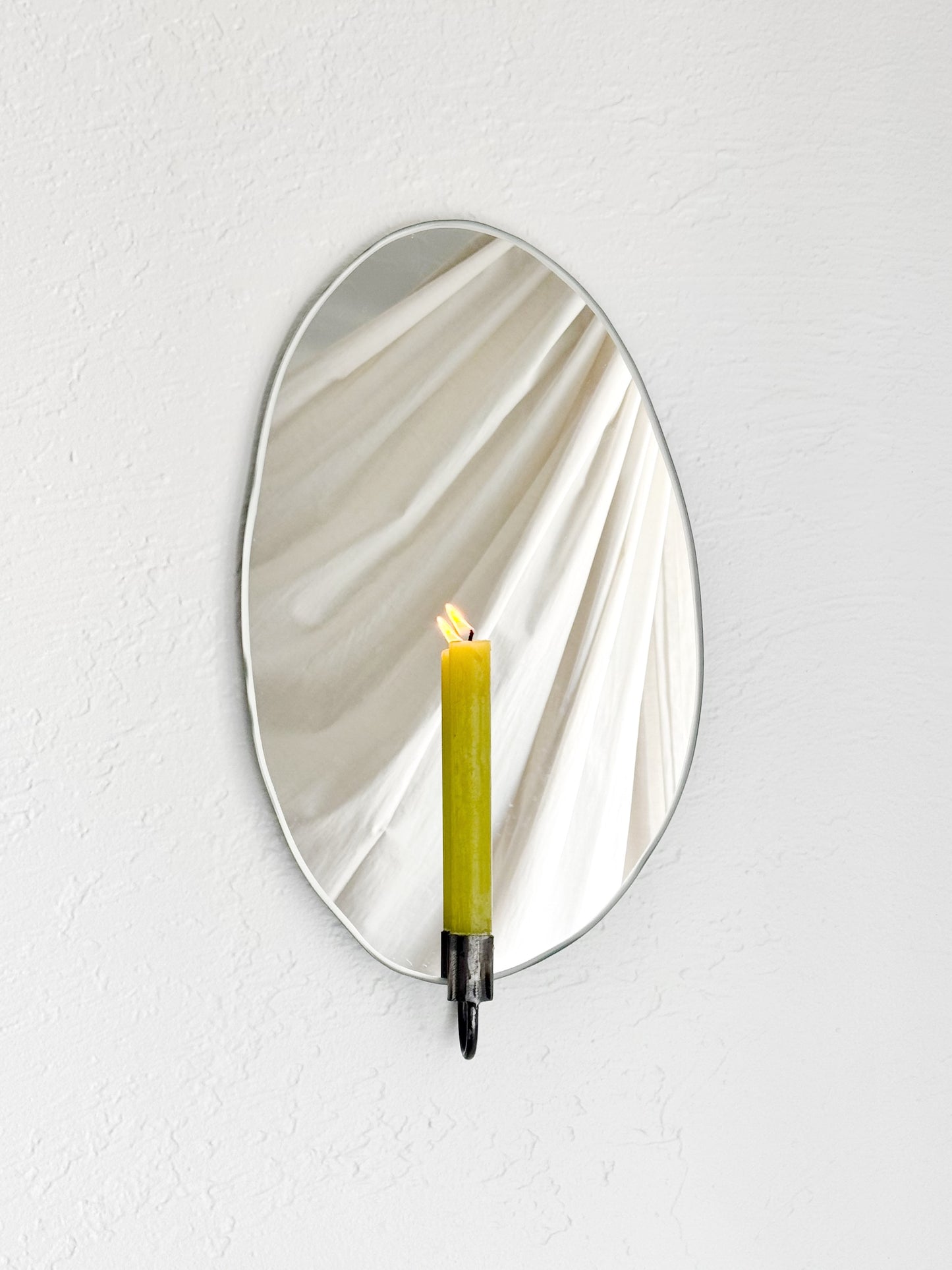 Straight view of the Mirror Candle Sconce or Mirror Candle Holder, wall mounted, with antique raw steel or silver candleholder and a green lit candle. The reflection shows the steel candle holder, light backdrop, candle & flame.