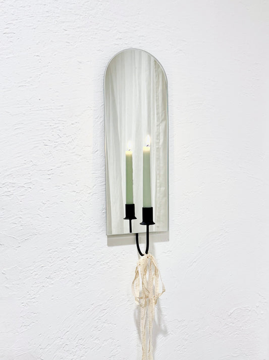 A frameless arch mirror on a white wall, wall mounted with a black candle holder sconce with a light green candle lit. The mirror is reflecting the candle flame and the candle holder has lacy ribbons tied to the bottom to give it a whimsical and feminine aesthetic.