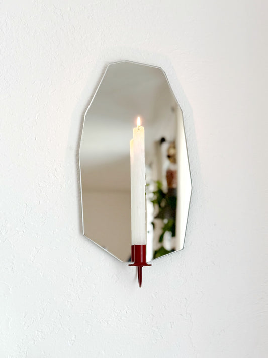 Gem Cut shaped Mirror Candle Sconce or Mirror Candle Holder, wall mounted. The candleholder is Cherry red and holding a white lit candle. The reflection is out of focus of some plants and cozy home decor.