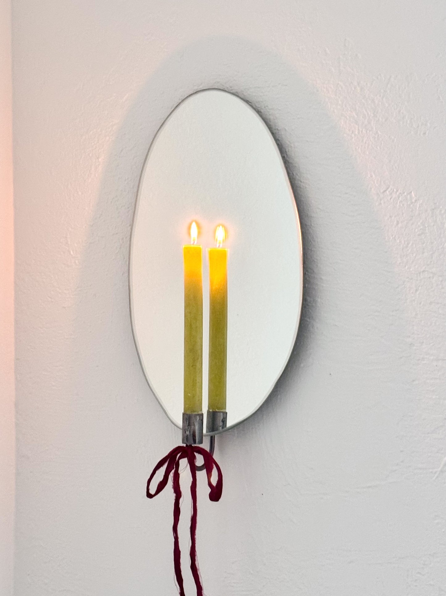 Angle view of the Mirror Candle Sconce or Mirror Candle Holder, wall mounted, with antique raw steel or silver candleholder & green lit candle. Red ribbon is tied into a bow on base of candleholder to give it a cozy and feminine aesthetic. Reflection shows steel candle holder, candle & flame.