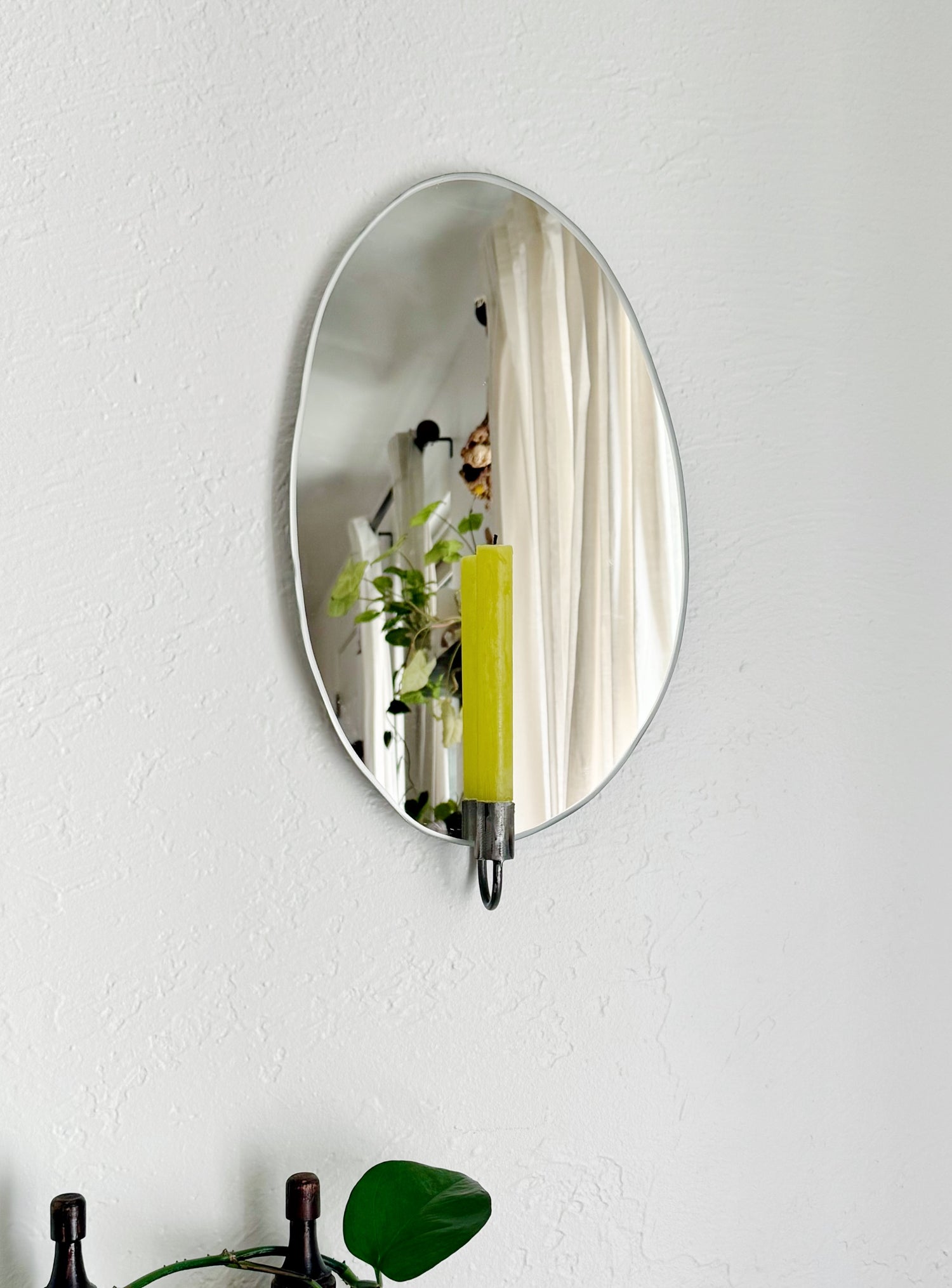 Straight view of the Mirror Candle Sconce or Mirror Candle Holder, wall mounted, with antique raw steel or silver candleholder and a green candle. The reflection shows the setting of a cozy home with plants and cozy room decor.