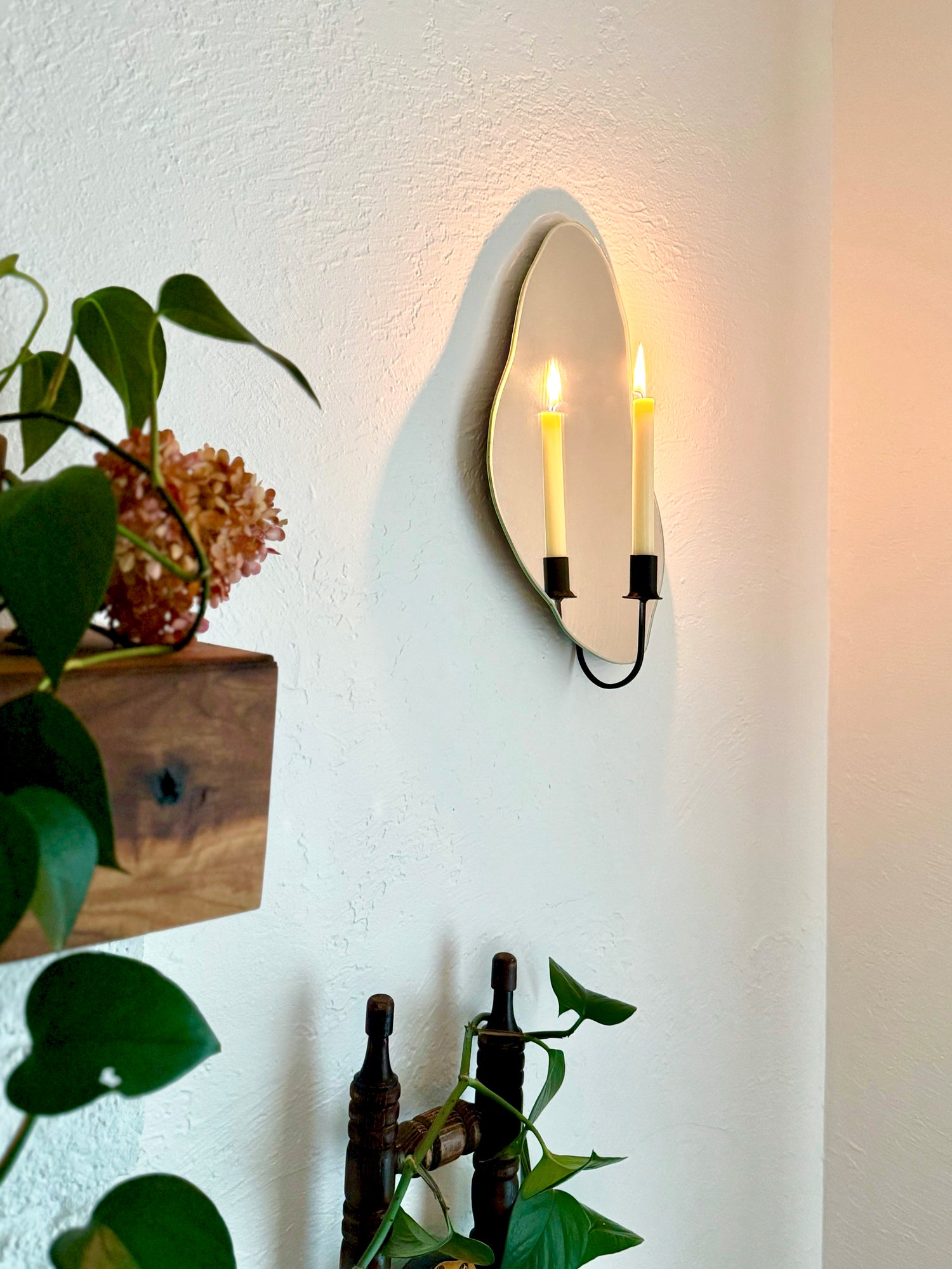 Side angle of organic shaped mirror candle sconce or mirror candle holder in a cozy home setting. The candle holder is black and wall mounted to a wall that is reflecting the warm light from the cream colored lit candle, creating a warm ambiance.