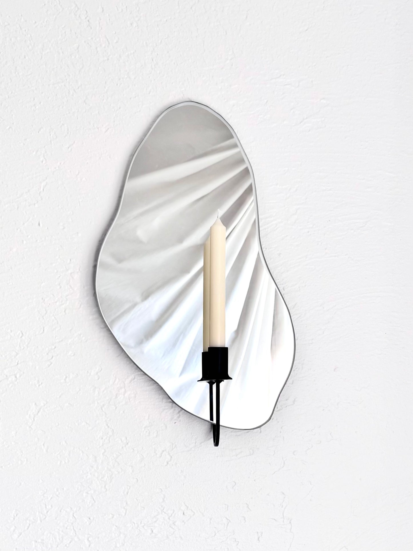 Front angle of the Shell Mirror Candle Sconce or Mirror Candle Holder, with a black candleholder and a cream colored unlit candle. The mirror candle holder is wall mounted on a white wall with a light colored backdrop reflected in the mirror.