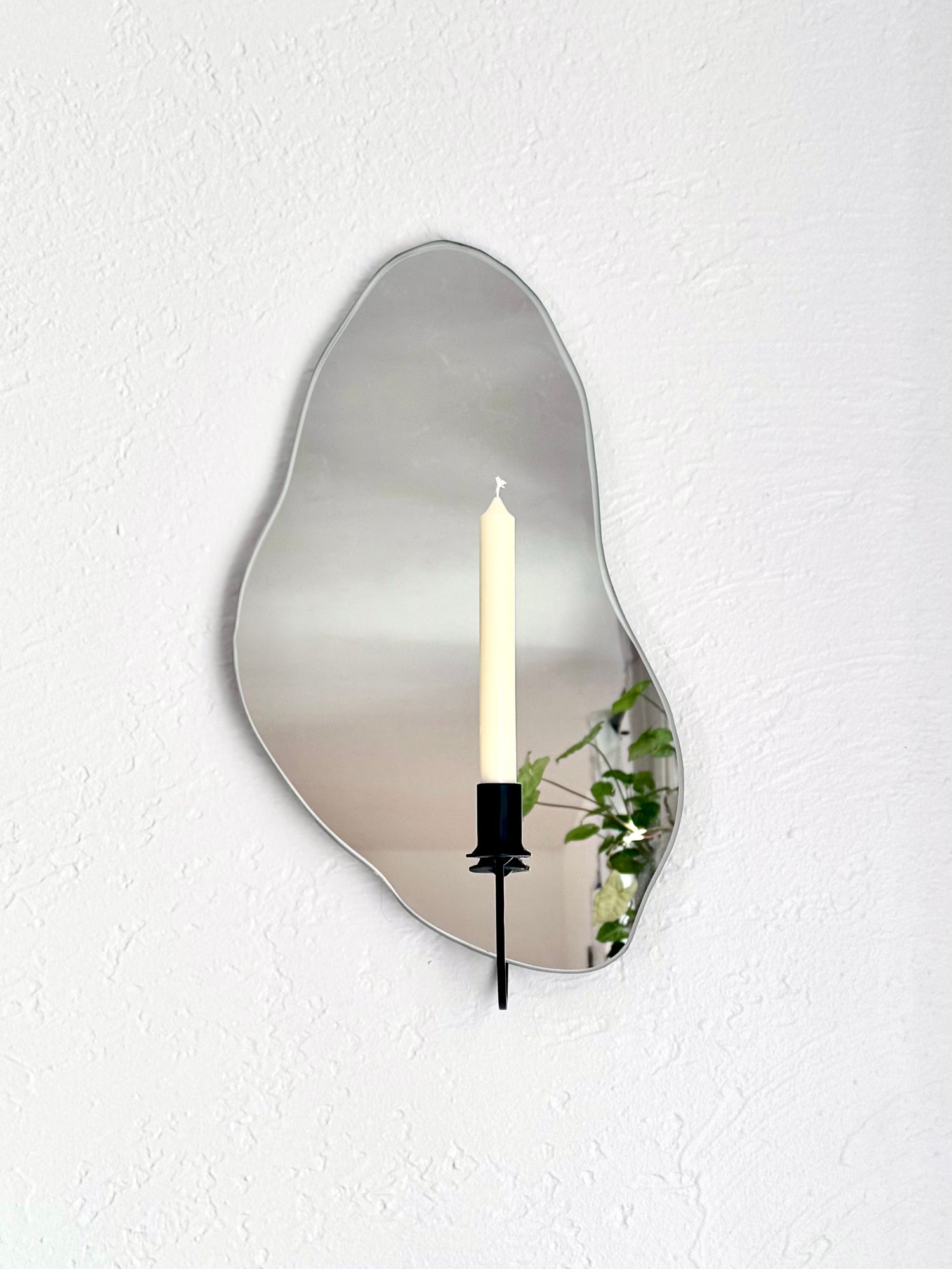 Front angle of the mirror candle sconce or mirror candle holder, with a black candle holder and a cream colored unlit candle. The mirror candle holder is wall mounted on a white wall with a light colored backdrop with plants reflected in the mirror.