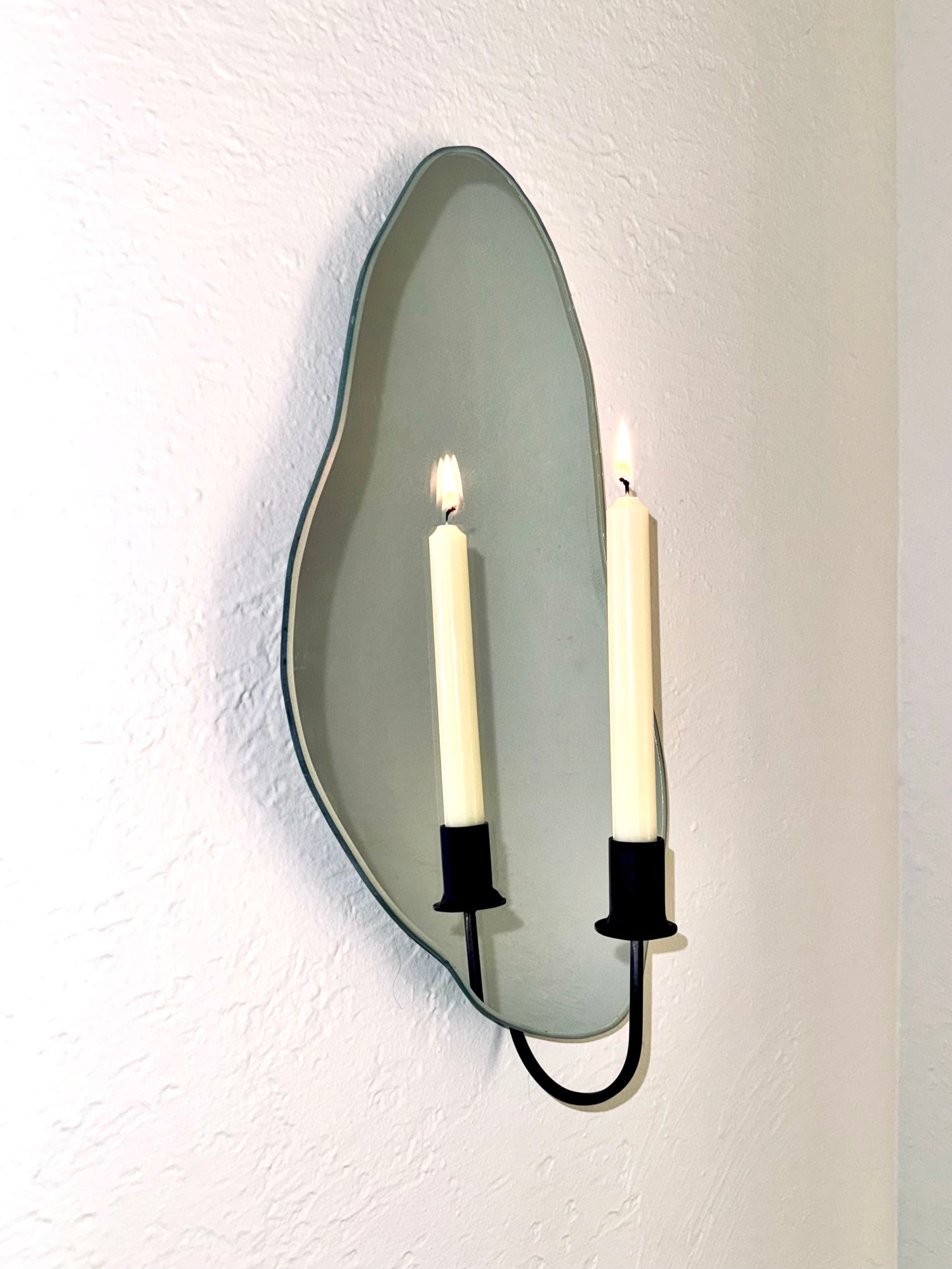 Side angle of mirror candle sconce or mirror candle holder with flash. The candleholder is black holding a cream colored lit candle. The flame and candle are reflected in the mirror. The mirror candle sconce is wall mounted to a white wall.