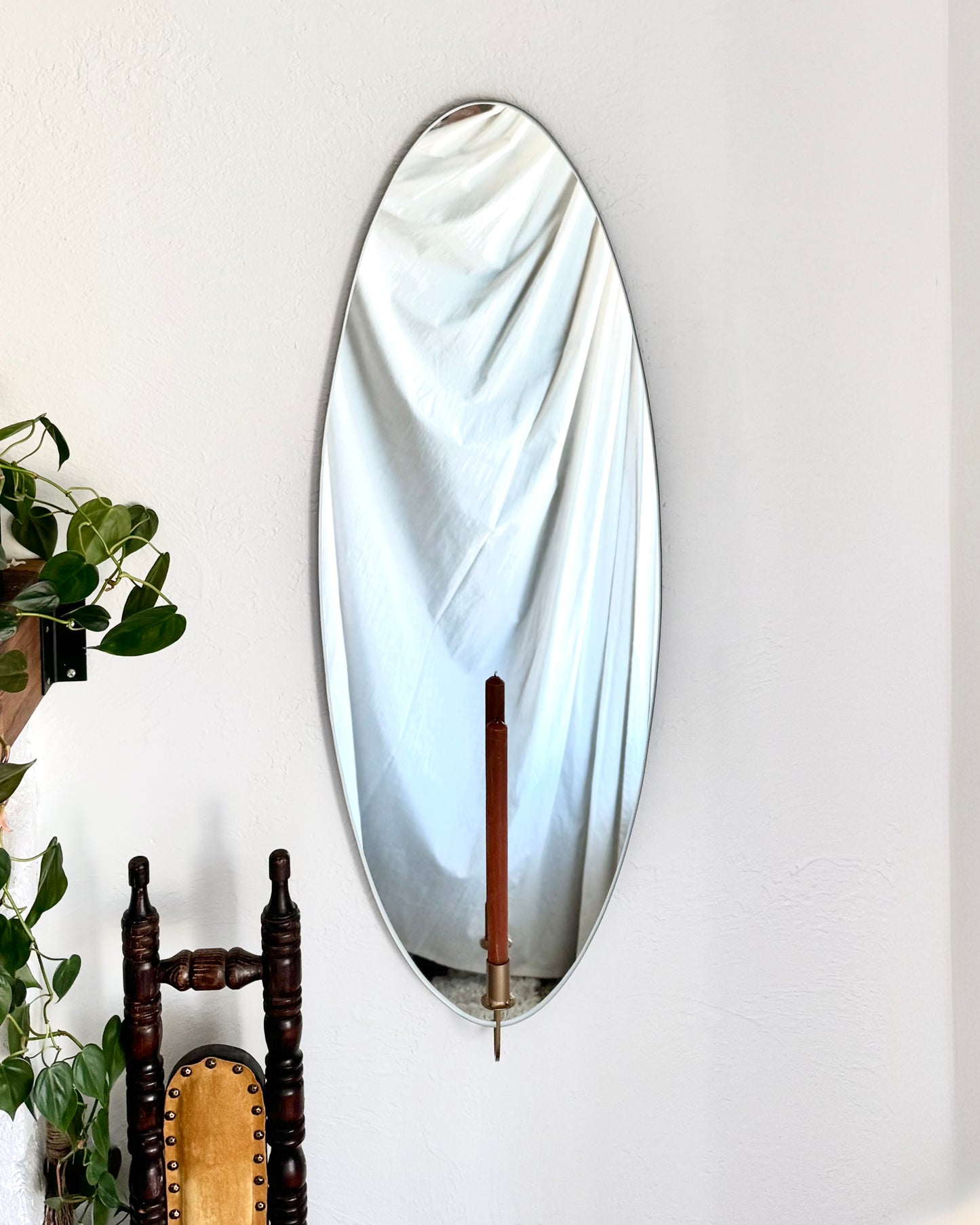 Large/Medium frameless wall mounted mirror with a candle sconce holding an unlit candle. The candleholder is gold with a brown unlit candle. There is an ivy plant next to the mirror and a vintage chair showing cozy home decor.