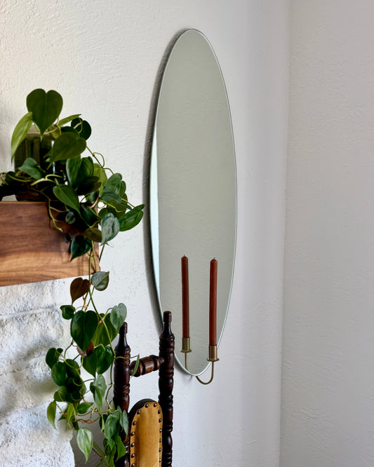 Medium/Large frameless wall mounted mirror with a candle sconce holding an unlit brown candle. The candleholder is gold and reflected in the mirror with the candle. There is an ivy plant next to the mirror and a vintage chair showing cozy home decor.