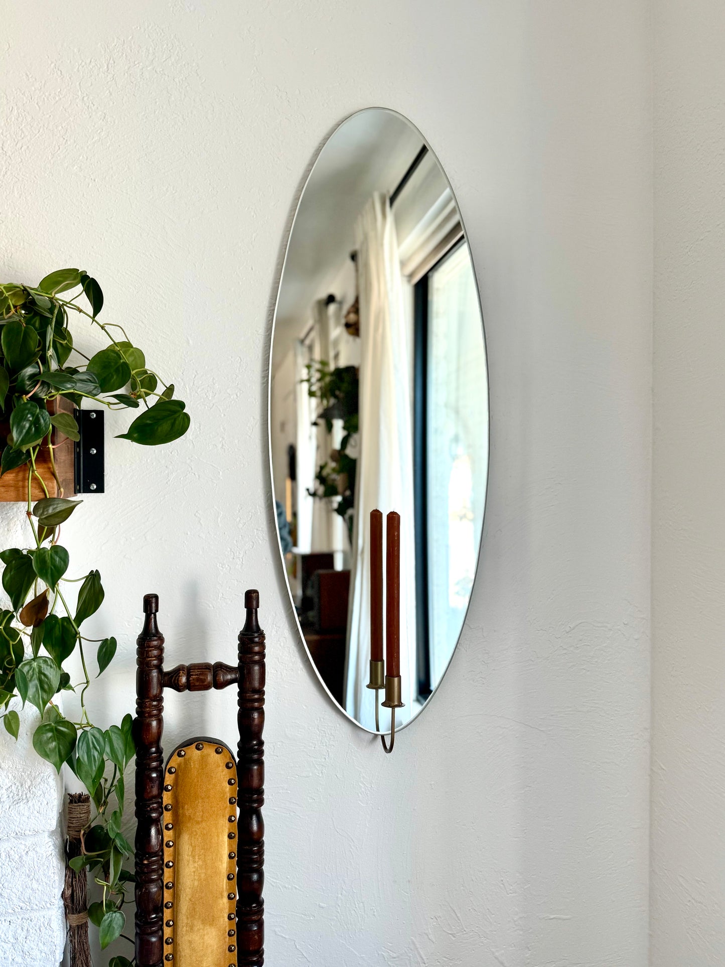 Large/Medium frameless wall mounted mirror with a candle sconce holding an unlit candle. The candleholder is gold and reflected in the mirror with the brown candle. There is an ivy plant next to the mirror and a vintage chair showing cozy home decor.