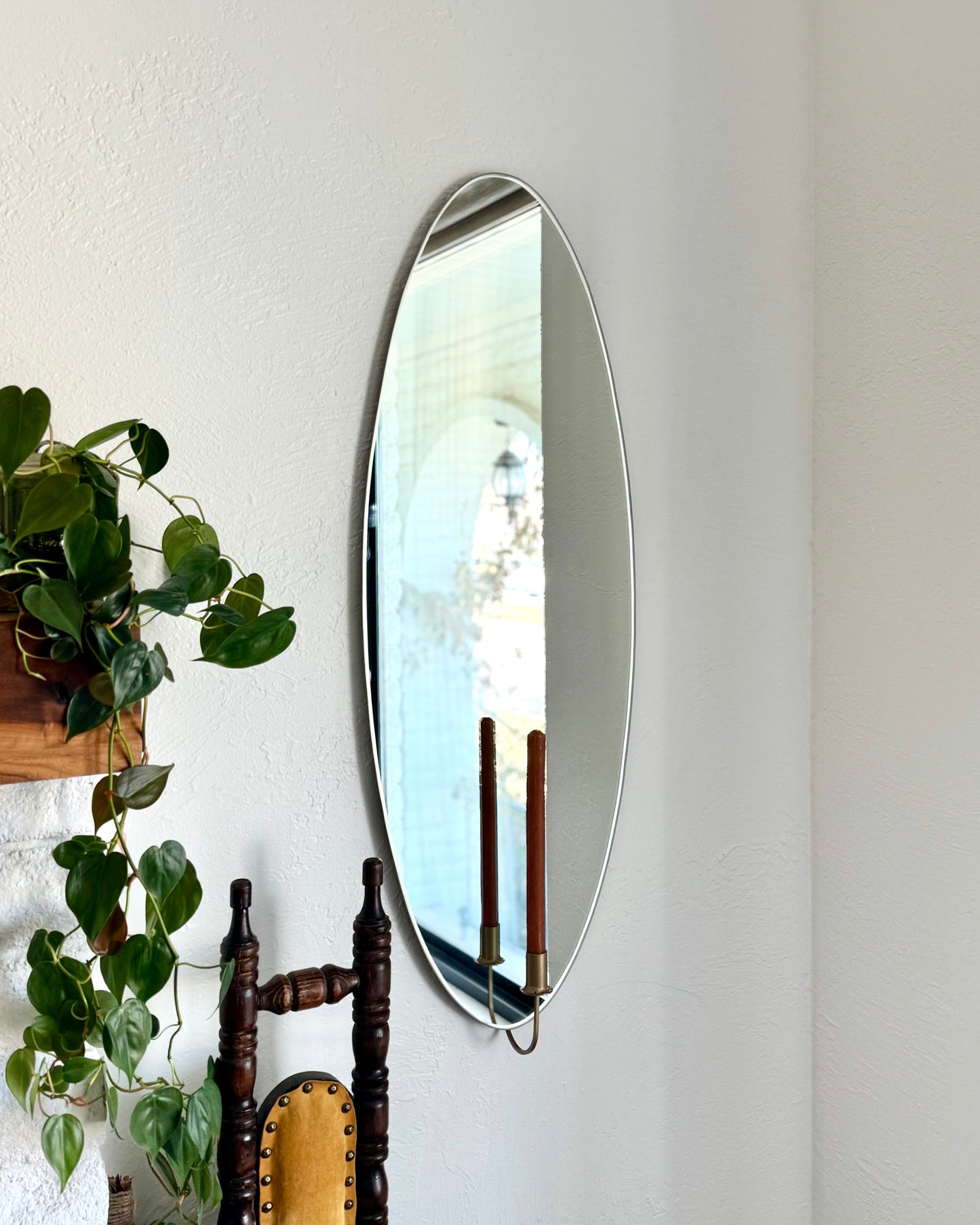 Large/Medium frameless wall mounted mirror with a candle sconce holding an unlit candle. The candleholder is gold and reflected in the mirror with the brown candle. There is an ivy plant next to the mirror and a vintage chair showing cozy home decor.