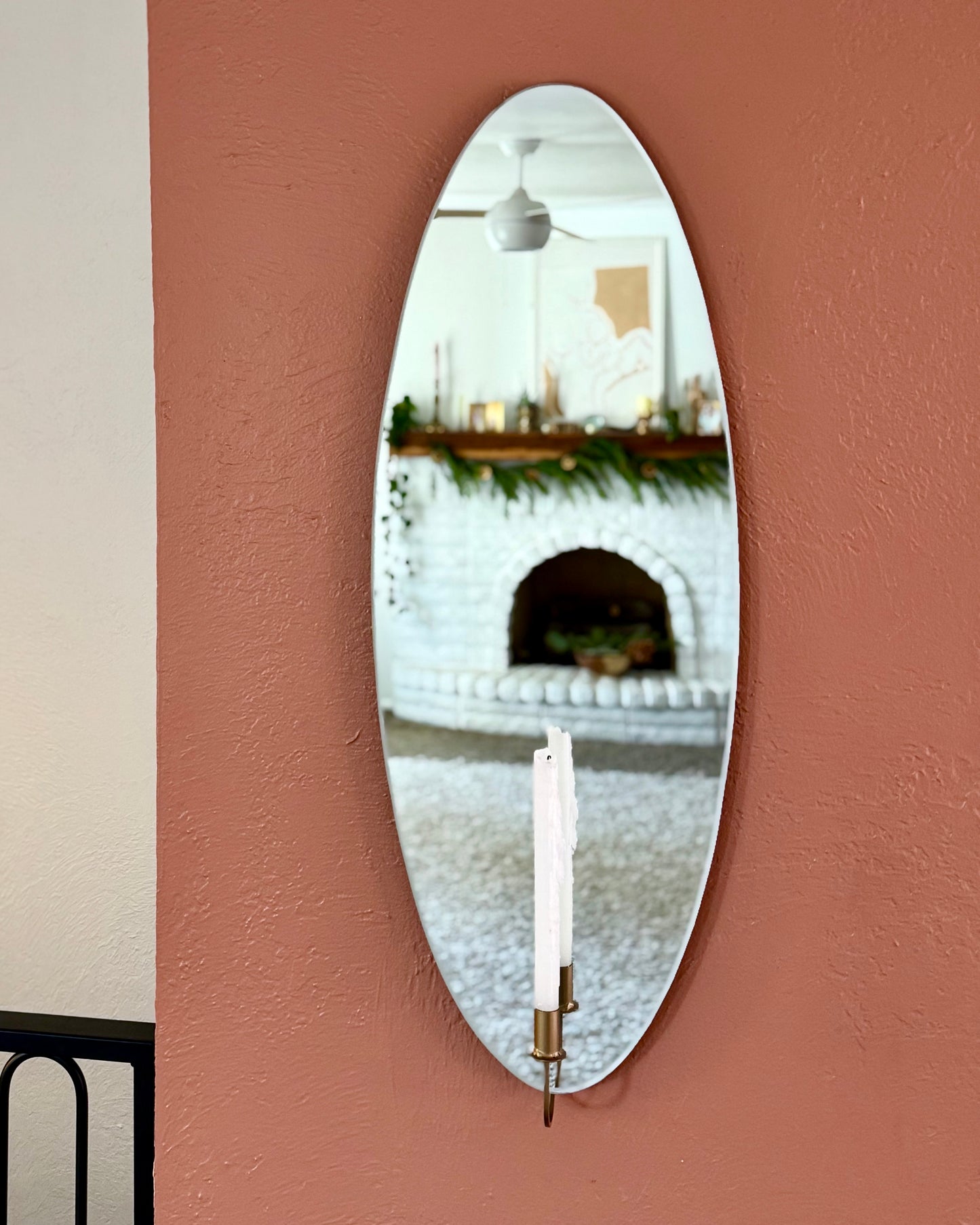 Medium/Large wall mounted frameless mirror candle sconce or mirror candle holder on a terracotta painted wall. The gold candleholder is holding a white unlit candle. The reflection is of an out-of-focus white brick fireplace giving cozy home vibes.