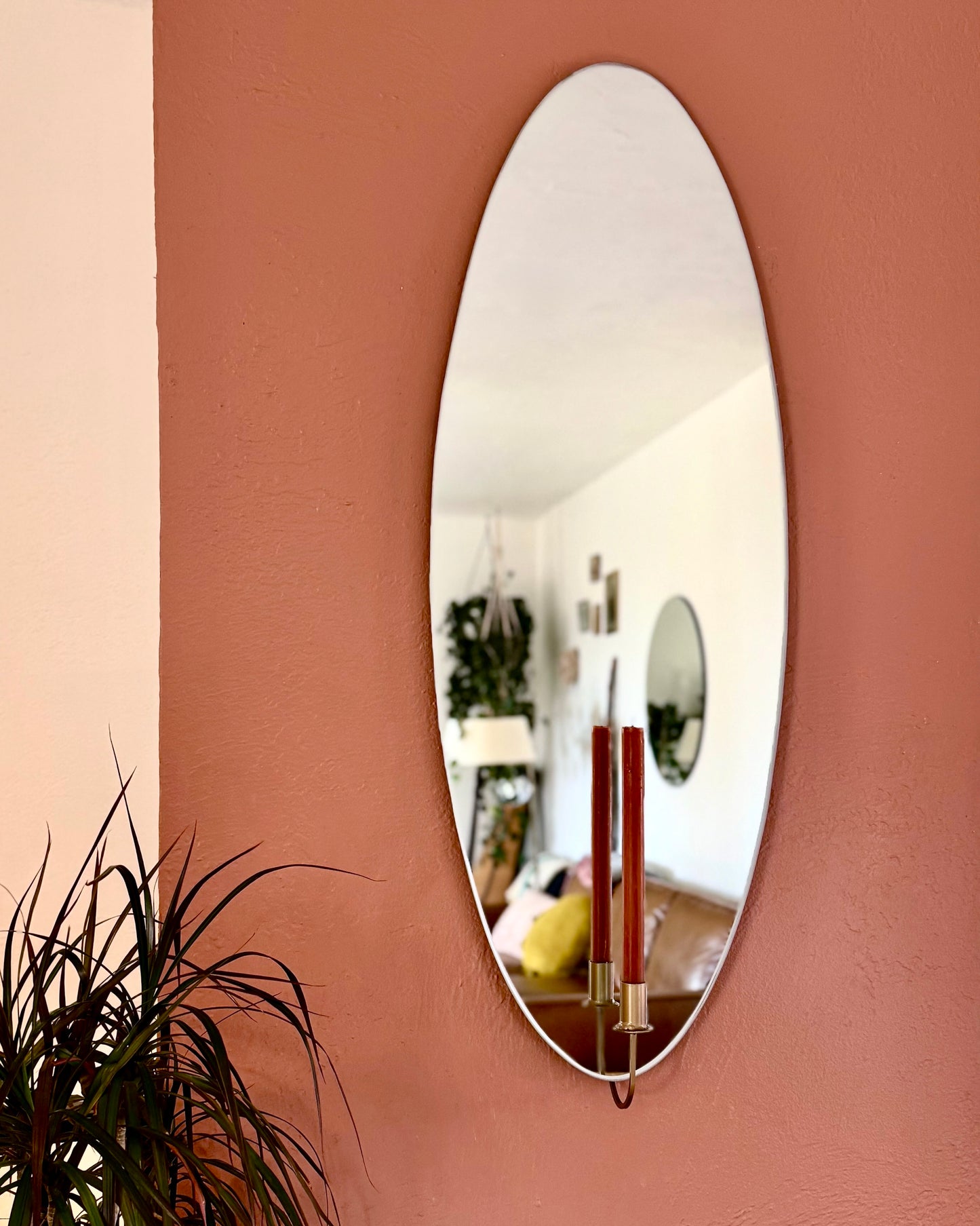 Large/Medium wall mounted frameless mirror candle sconce or mirror candle holder on a terracotta painted wall. The gold candleholder is holding a brown unlit candle. The reflection is of the candle & candle sconce & out-of-focus cozy home decor.