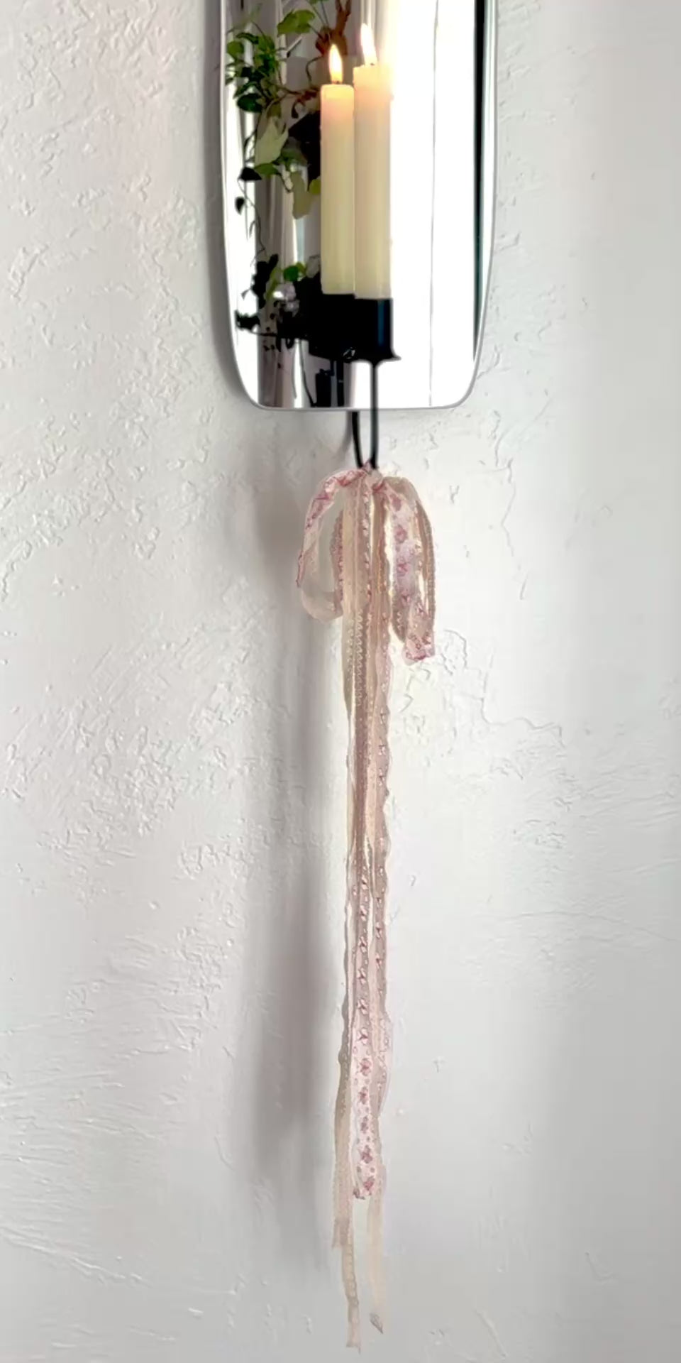 Video of the Oar Mirror Candle Sconce or Mirror Candle Holder, wall mounted. The black candleholder has lacy pink & cream ribbons tied to bottom to create a whimsical aesthetic. Reflection is of the candle holder & a cream lit candle with flame, plants & cozy decor.