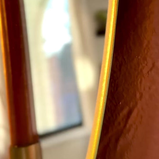 Video of Large/Medium wall mounted frameless mirror candle sconce or mirror candle holder on a terracotta painted wall. The gold candleholder is holding a brown unlit candle. The reflection is of the candle & candle sconce & out-of-focus cozy home decor. The sunset is casting a warm light and shadow onto the mirror sconce.