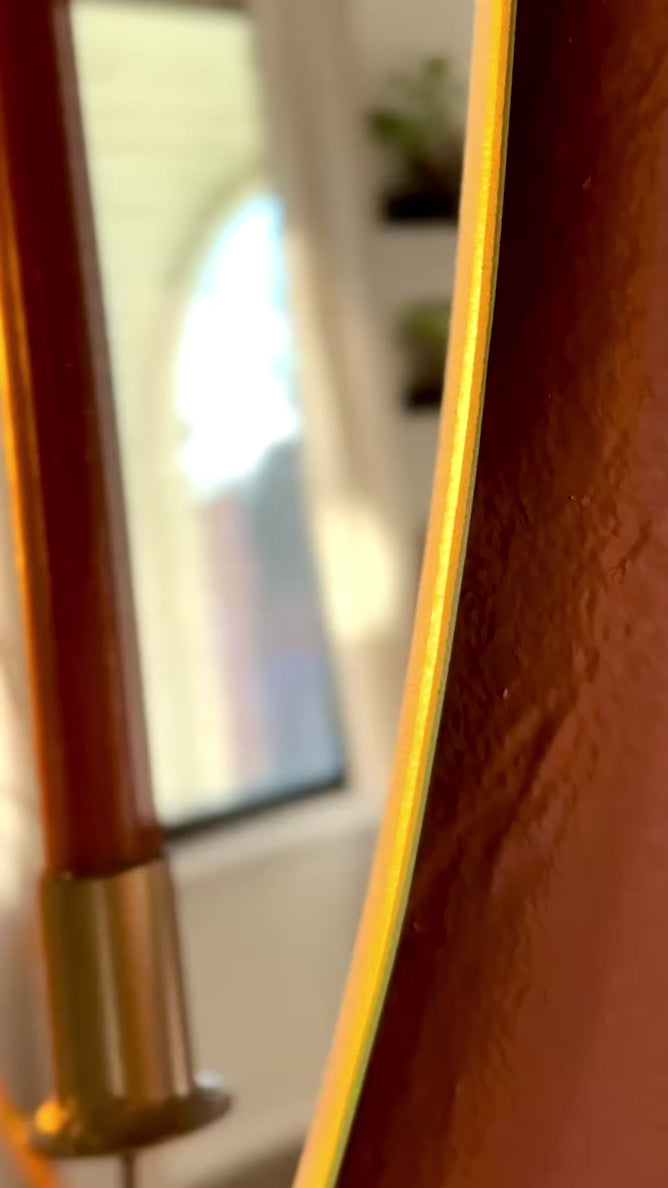 Video of Large/Medium wall mounted frameless mirror candle sconce or mirror candle holder on a terracotta painted wall. The gold candleholder is holding a brown unlit candle. The reflection is of the candle & candle sconce & out-of-focus cozy home decor. The sunset is casting a warm light and shadow onto the mirror sconce.