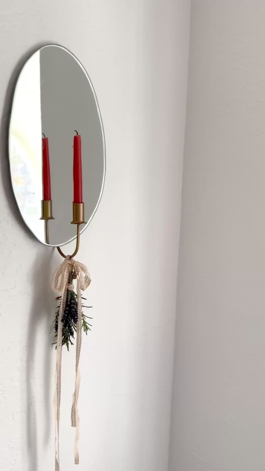 Video of the Mirror Candle Sconce or Mirror Candle Holder, wall mounted, with the gold candleholder and a red candle. The base of the gold candle holder has cream ribbon tied into a bow with a small bundle of dried herbs (rosemary & lavender) creating an inviting aesthetic. The reflection shows the light backdrop. A woman walks to the wall candle sconce and lights the candle.