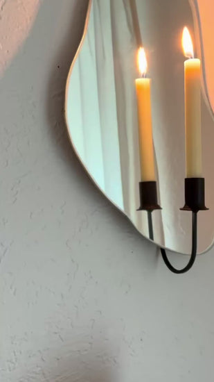 Video of the Shell Mirror Candle Sconce or Mirror Candle Holder with a black candleholder holding a cream colored lit candle. The video moves slowly from the side angle to more of a straight shot of the sconce, showing the warmth the lighting and mirror bring. Reflection in mirror is a cozy home setting with plants.